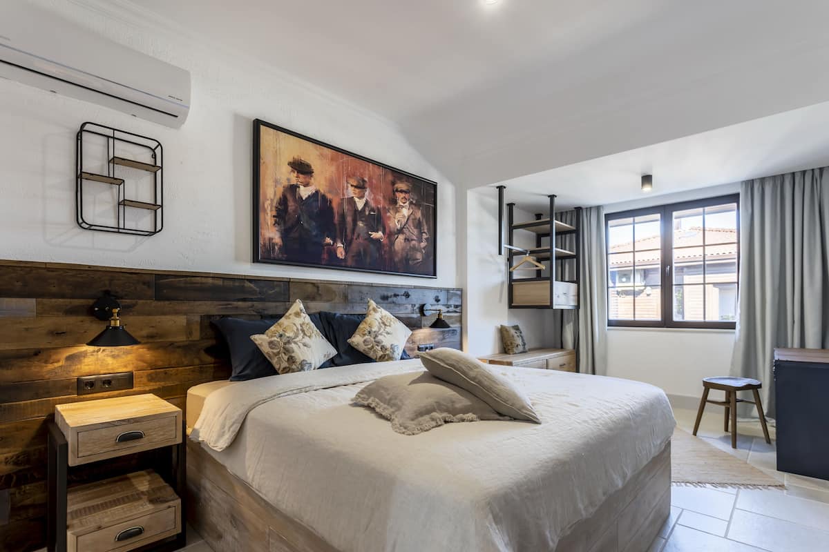 A modern bedroom with a wooden headboard, a large bed with multiple pillows, a bedside table with drawers, a wall-mounted shelf, and a large window with curtains.