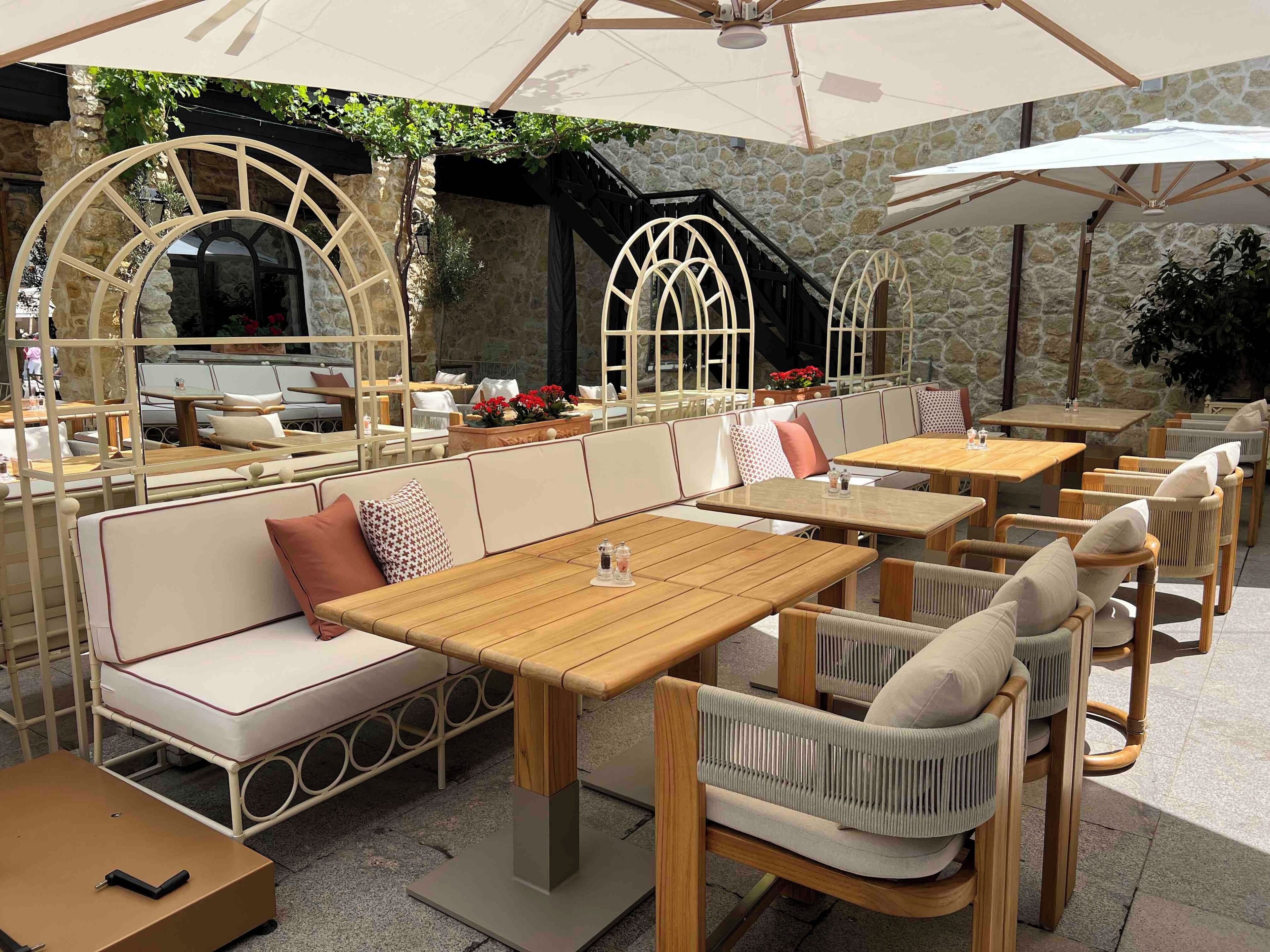 An outdoor seating area with wooden tables and cushioned benches, stone walls, and a covered section with umbrellas.