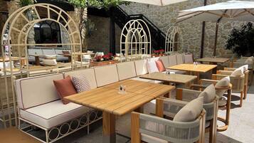 An outdoor seating area with wooden tables and cushioned benches, stone walls, and a covered section with umbrellas.