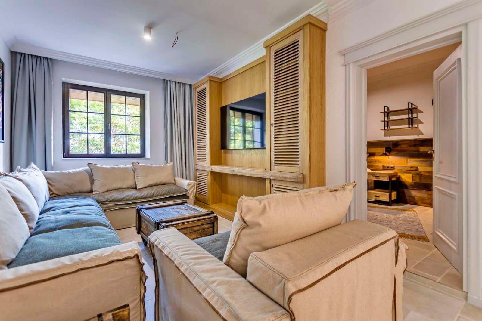A spacious living room with a large sofa set, a wooden cabinet with a television, and a view of the outdoors through a window.