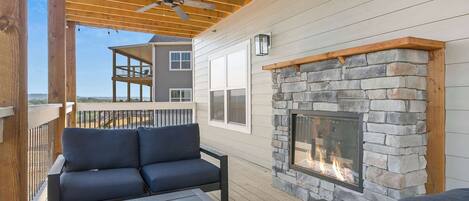 A covered outdoor deck with a stone fireplace, wooden furniture, and a view of a neighboring building.