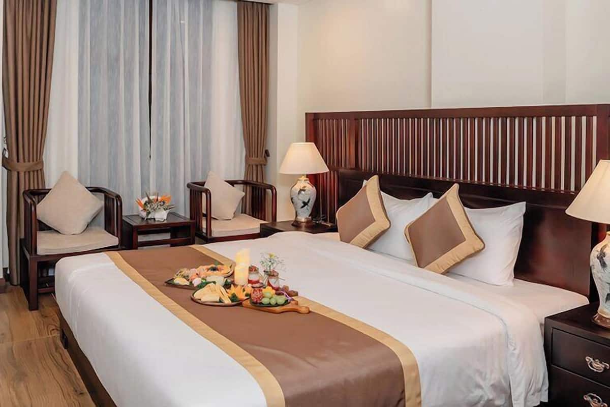 Une chambre d’hôtel avec un grand lit, deux tables de chevet avec des lampes, une tête de lit en bois, un bureau en bois, une chaise et une petite table agrémentée d’un arrangement floral.