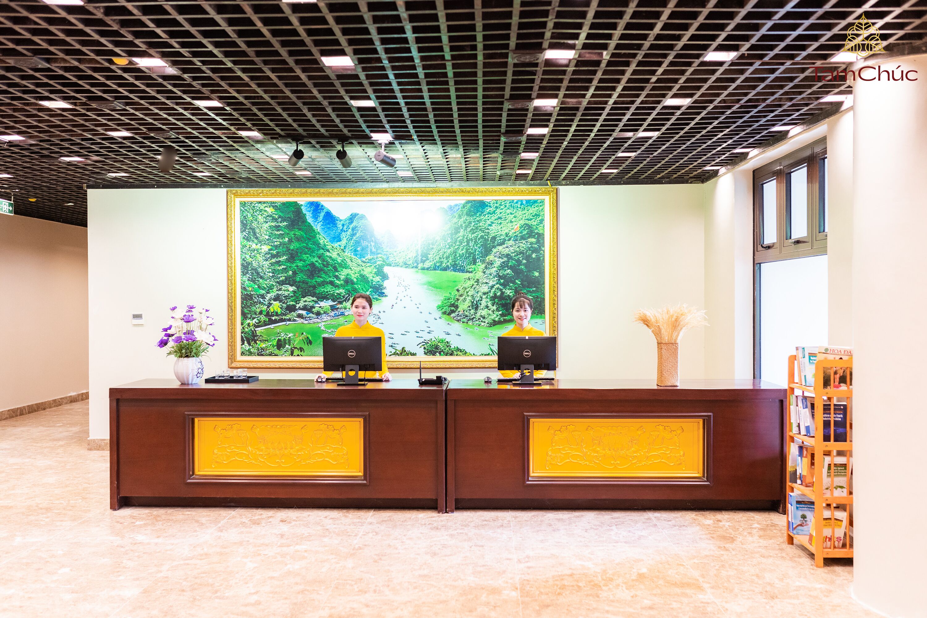 A reception desk with two monitors, a large framed picture of a landscape, and a bookshelf.