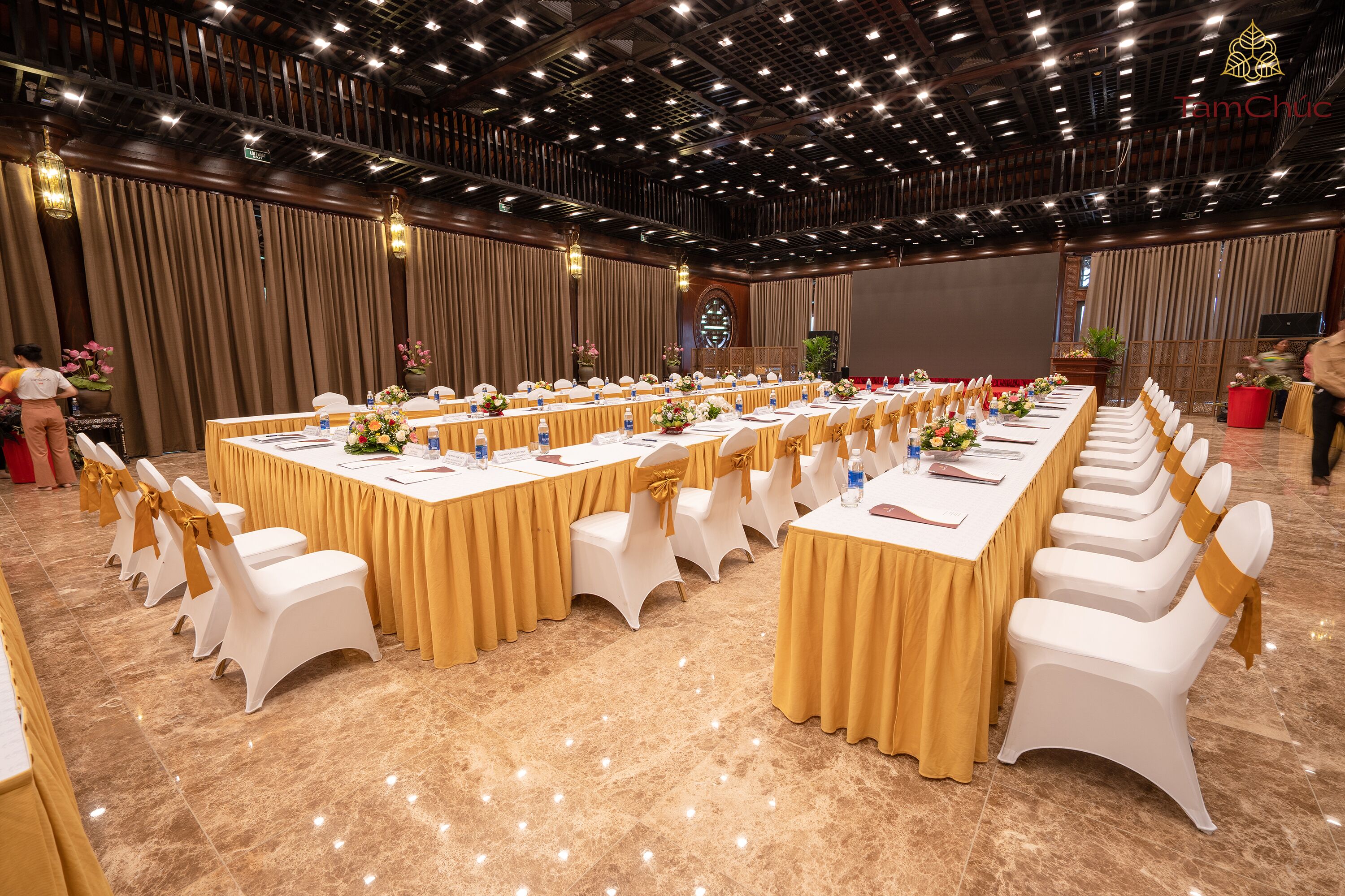 A banquet hall with long tables, white chairs, and gold drapes.
