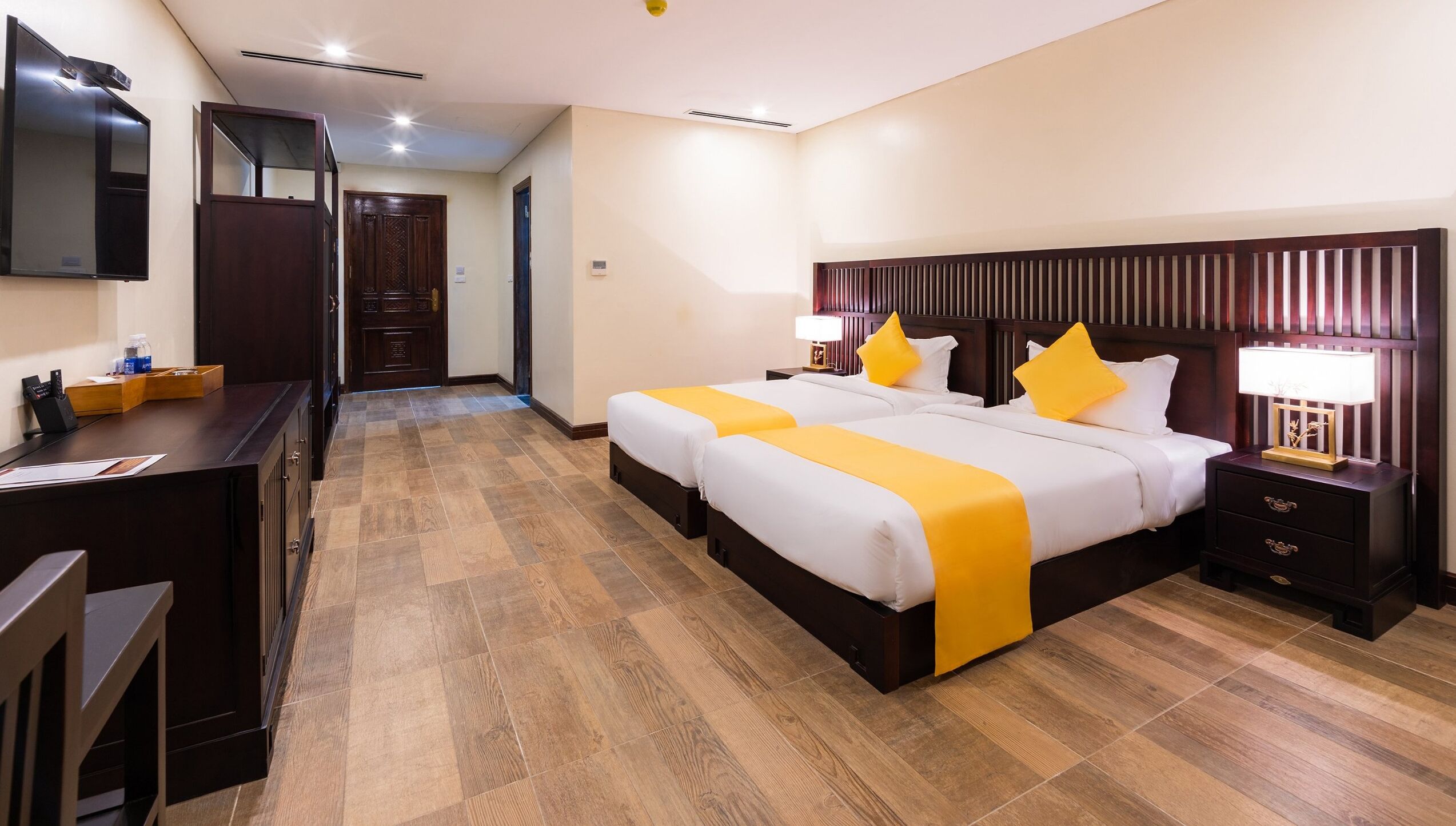 Habitación de hotel con suelo de madera, dos camas con ropa de cama blanca y amarilla, un escritorio con espejo y una mesita de noche con lámpara.