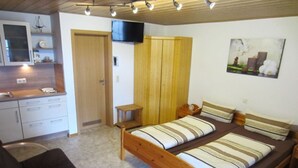 Un dormitorio con una cama de madera, una mesita de noche pequeña, una silla, un televisor montado en la pared y una zona de cocina con armarios y encimera.