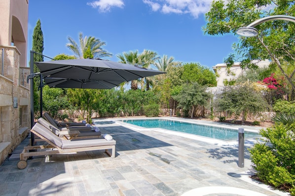 A pool area with lounge chairs and an umbrella, surrounded by greenery and a stone building.