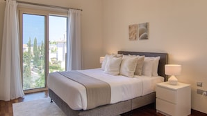 A bedroom with a bed, a nightstand, a window with curtains, and a view of the outside. - Phaedrus Living: Amorosa 13 Beachside Villa (Neo Chorio)