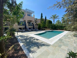 A pool area with lounge chairs and umbrellas, surrounded by trees and a building with a pergola.