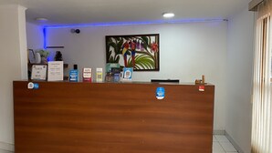 A hotel reception desk with a wooden counter, a framed tropical painting, and various informational signs. - Hotel Itavera (Puerto Iguazú)