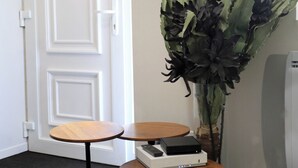 Une pièce avec une porte blanche, un téléviseur à écran plat fixé au mur, un grand vase contenant des fleurs foncées et une petite table sur laquelle se trouve un haut-parleur.