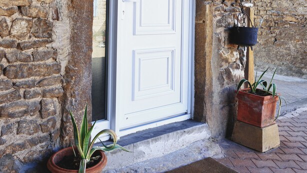 Une porte blanche ornée d’un panneau décoratif, un pot de fleurs et un paillasson devant une entrée pavée de briques.