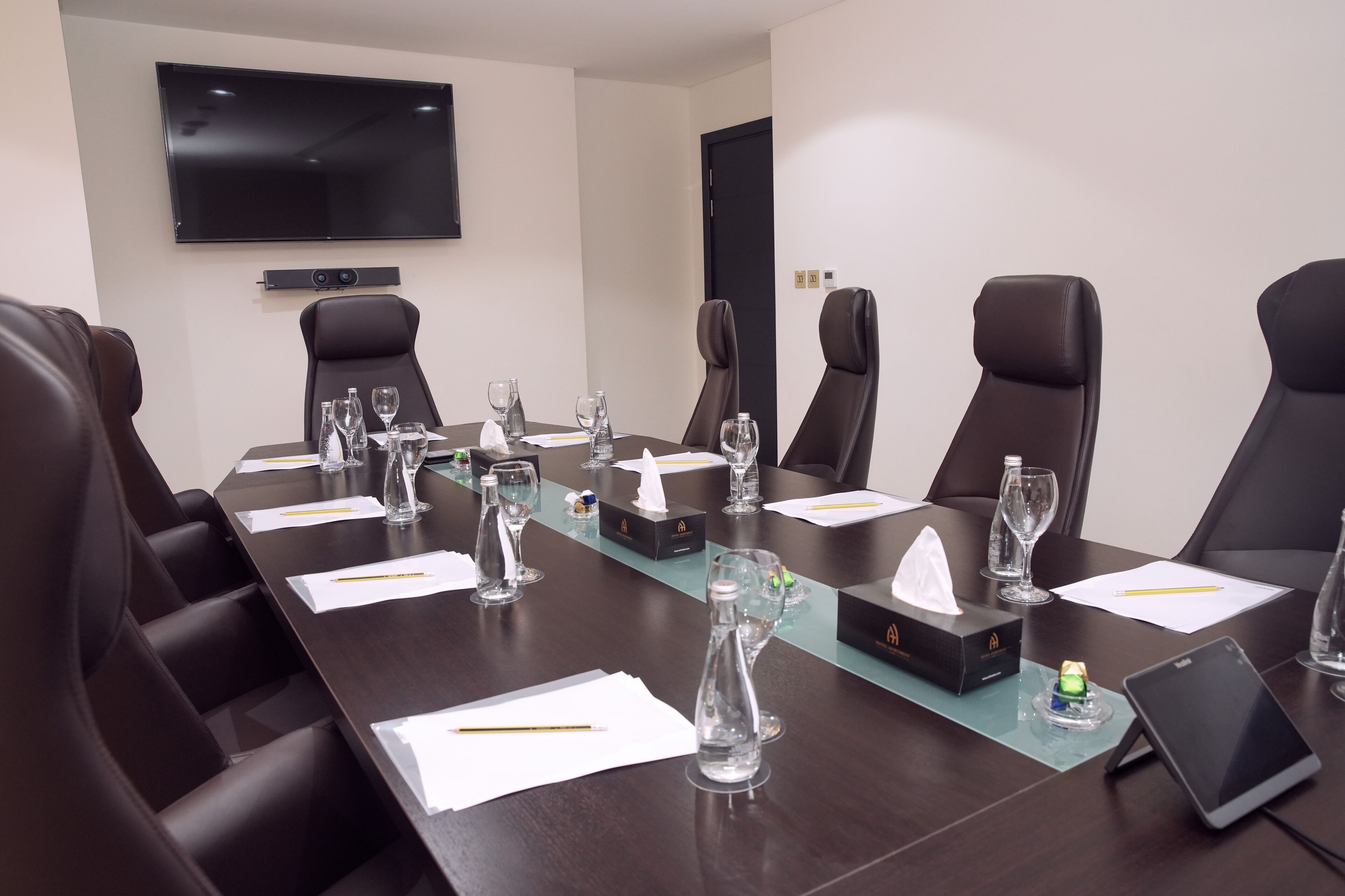 A conference room with a long table, black leather chairs, a flat-screen TV, and various items like glasses, tissues, and a tablet.