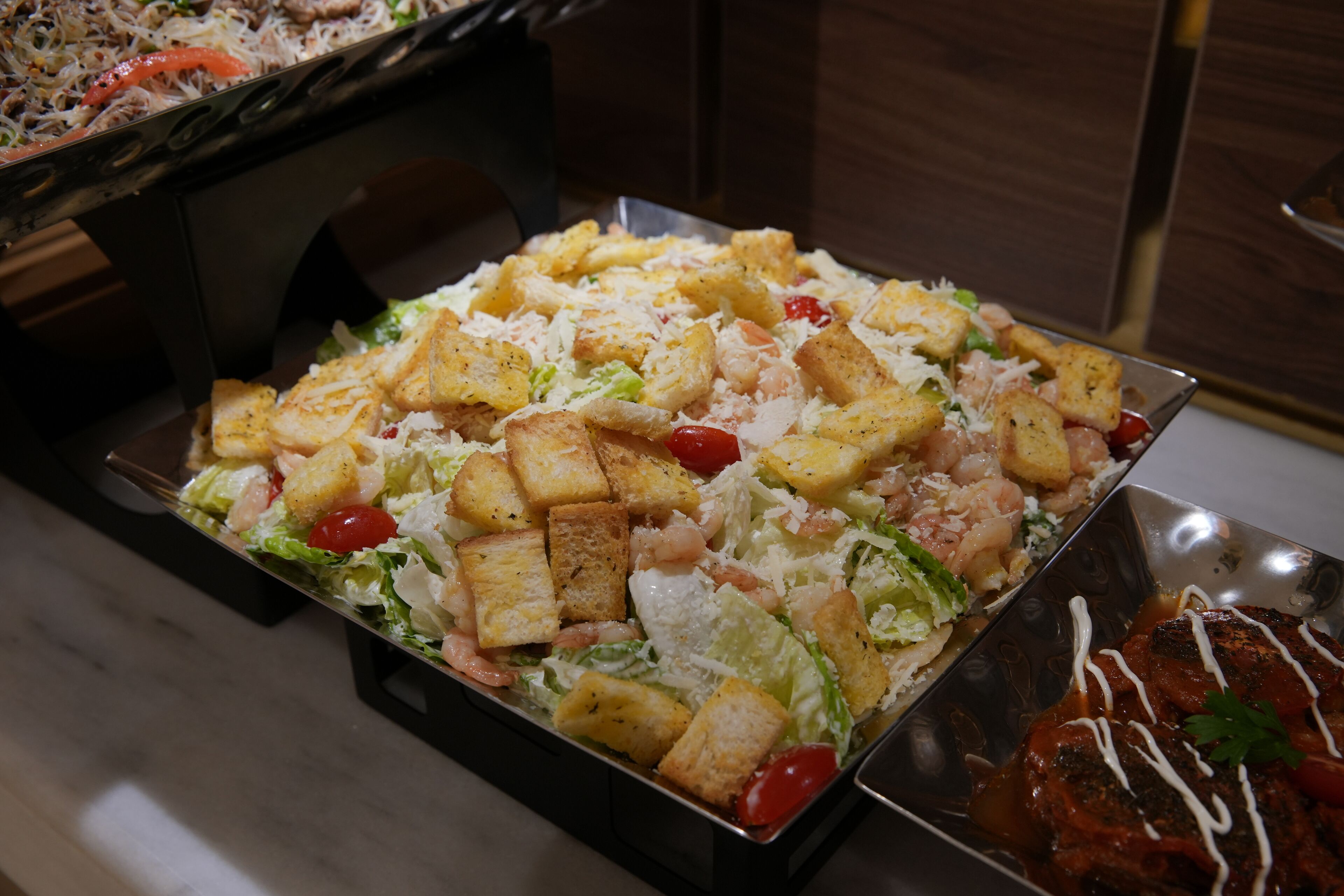 Un bufé con diversos platos, incluyendo una ensalada con crutones y queso rallado, y un plato de carne con salsa.