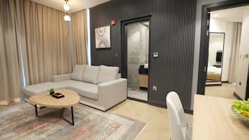 A modern hotel room with a sofa, a round coffee table, a dining table with fruit, and a large mirror.