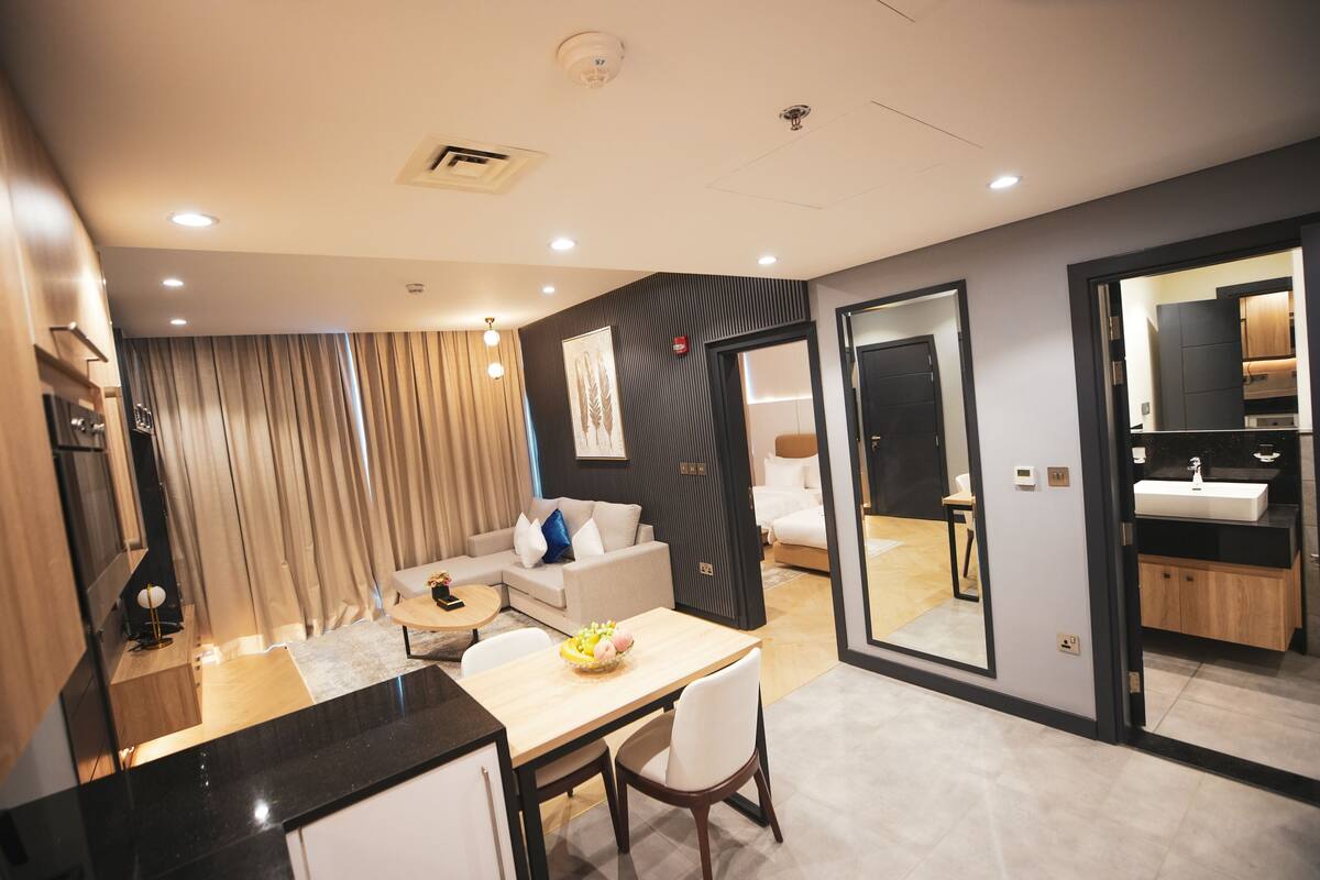 Ein modernes Hotelzimmer mit Essbereich, einer Sitzecke, einem kleinen Tisch mit Obstschale und einem großen Spiegel.