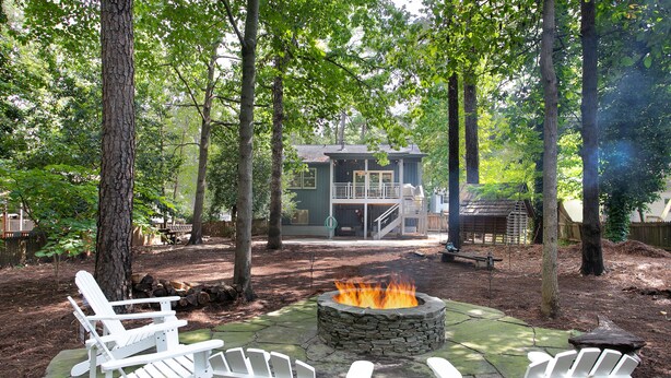 Ein Steinkamin wird von weißen Adirondack-Stühlen in einem bewaldeten Garten umgeben, im Hintergrund ist ein Haus zu sehen.