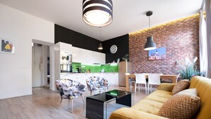 Uma sala de estar moderna com um sofá amarelo, uma mesa de centro de vidro e cadeiras com padrão floral. Uma cozinha com painel verde e parede de tijolos é visível ao fundo.