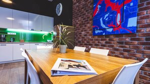 Cozinha moderna com armários brancos, bancada iluminada em verde e parede de tijolos com uma grande pintura abstrata.