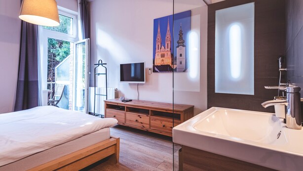 Quarto moderno com cama grande, cômoda de madeira, televisão e banheiro com espelho amplo e pia.