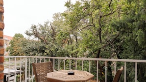 Um deck de madeira com mesa e cadeiras redondas, cercado por um corrimão branco, com vista para um jardim com árvores e um muro de tijolos.