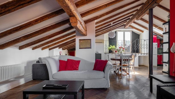 A spacious loft with exposed wooden beams, a grey sofa with red cushions, a black coffee table, and a view of the kitchen and balcony.