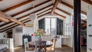 A spacious loft with exposed wooden beams, a dining area with a round table and chairs, and a view of the kitchen through a glass door.