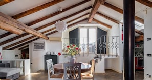 A spacious loft with exposed wooden beams, a dining area with a round table and chairs, and a view of the kitchen through a glass door.