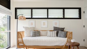 Un sofá de ratán con cojines, una lámpara de pie y un taburete de madera en una habitación con ventanas amplias y obras de arte enmarcadas en la pared.