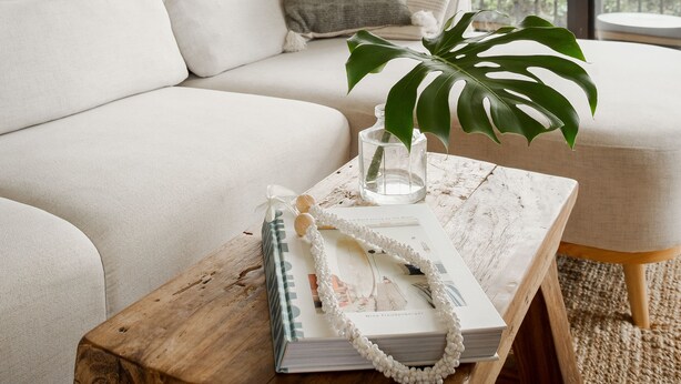 Una sala de estar moderna con un sofá blanco, una mesa de centro de madera con un libro y una planta, y un balcón con vista a árboles.