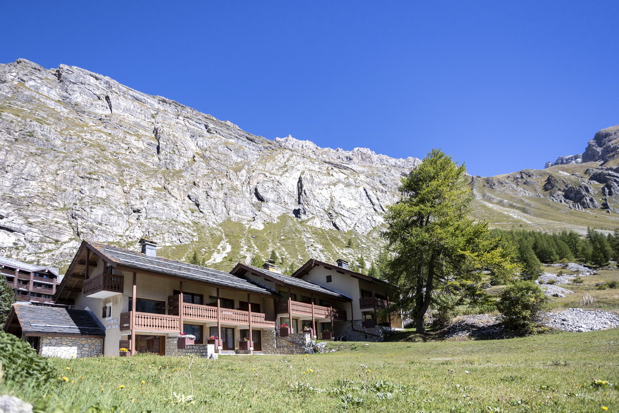 A mountain lodge with multiple balconies and a stone facade, set against a backdrop of rocky mountains and a clear sky.