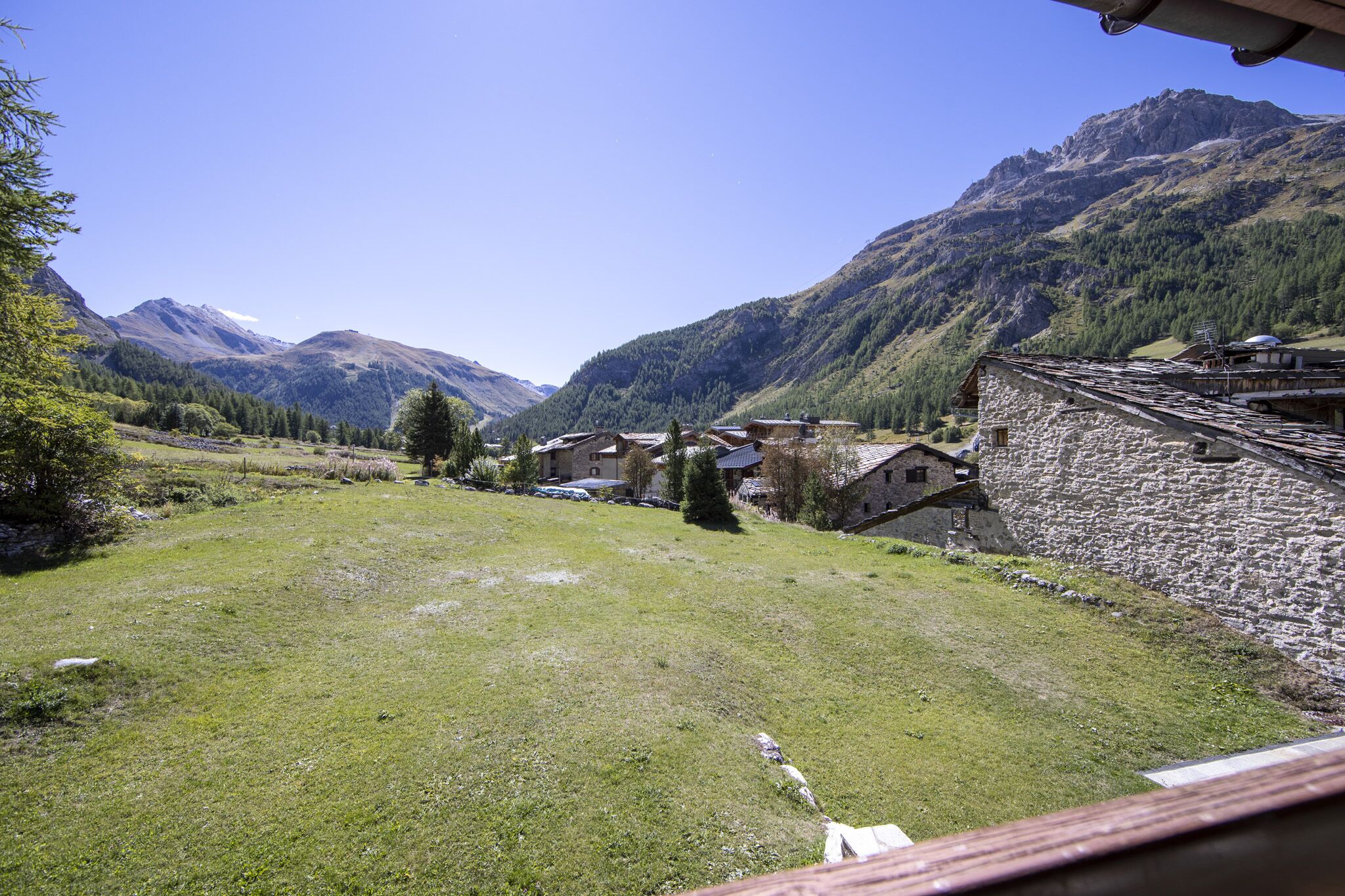 Un village rural avec des bâtiments en pierre, un champ verdoyant et des montagnes en arrière-plan.