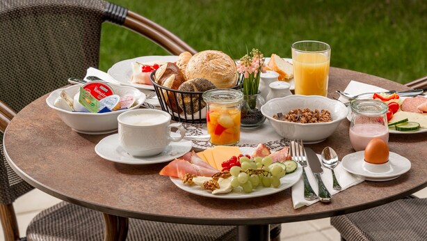 A round dining table with breakfast items, including bread, fruit, and beverages, set outdoors with wicker chairs. - Villa Elisa (Salzburg)