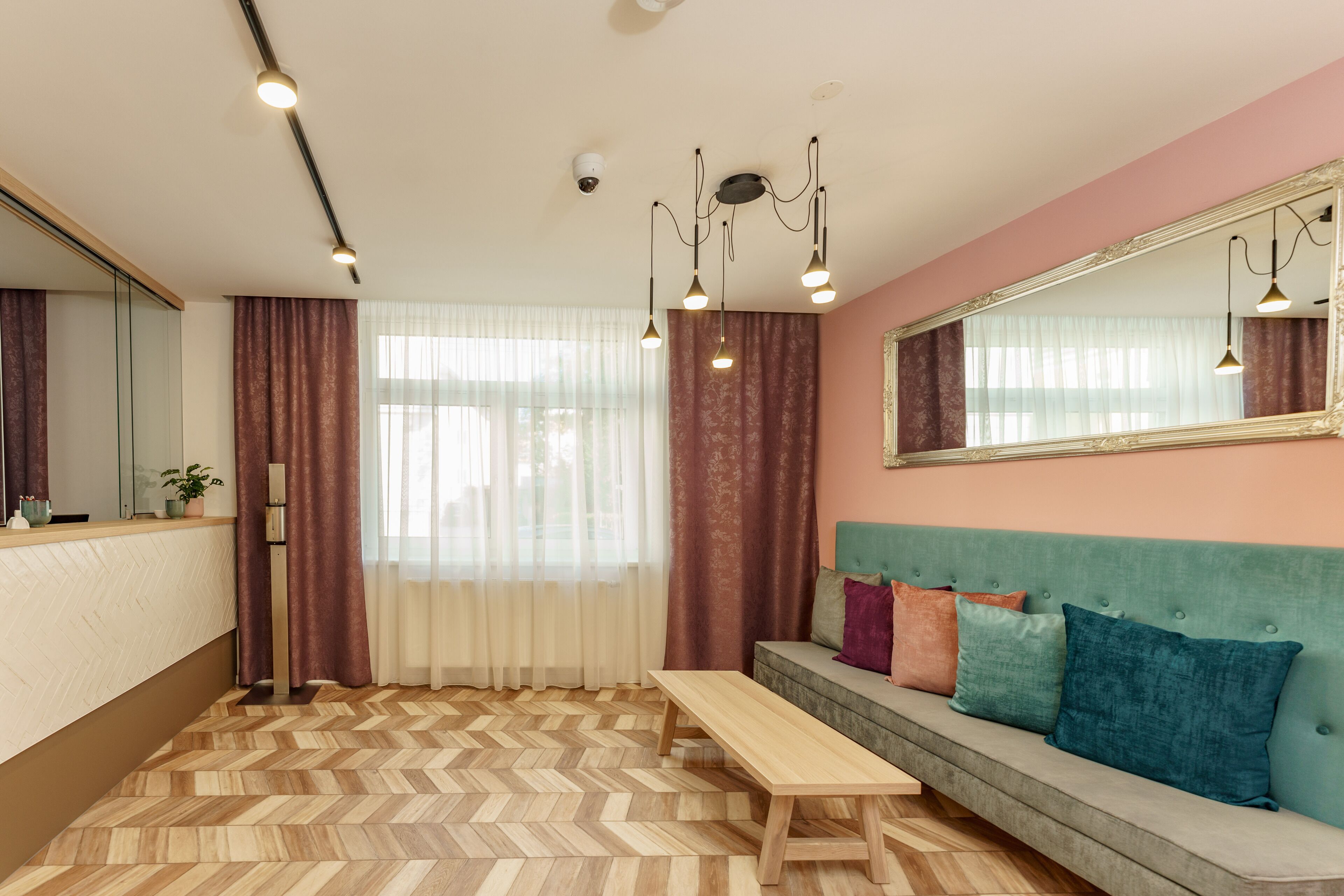 Quarto de hotel moderno com piso em padrão losango, um banco de madeira, um sofá verde-água com almofadas coloridas, um espelho grande e iluminação pendente.