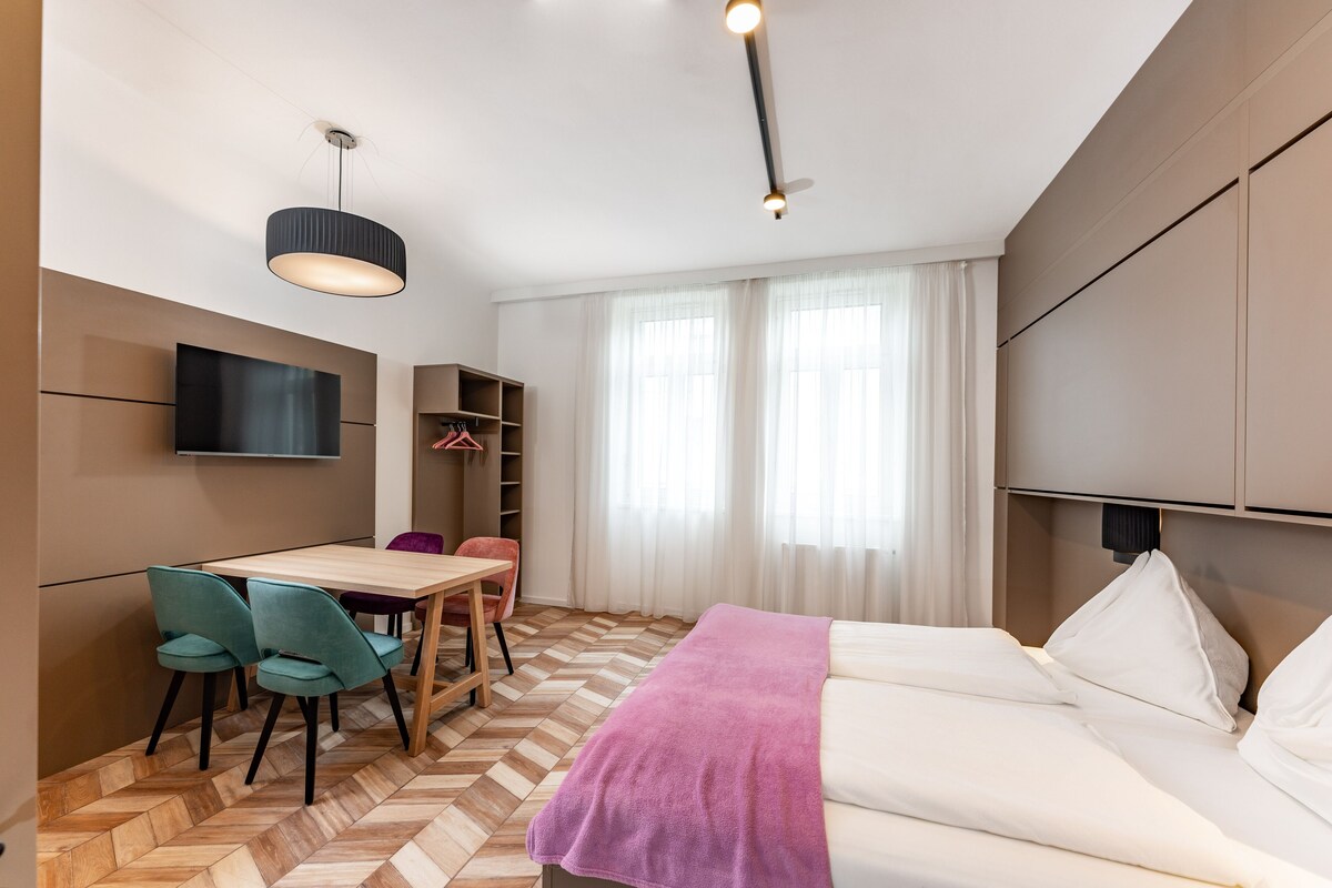 A modern hotel room with a wooden herringbone floor, a bed with white linens, a desk with teal chairs, a television mounted on the wall, and a wardrobe with hanging space.