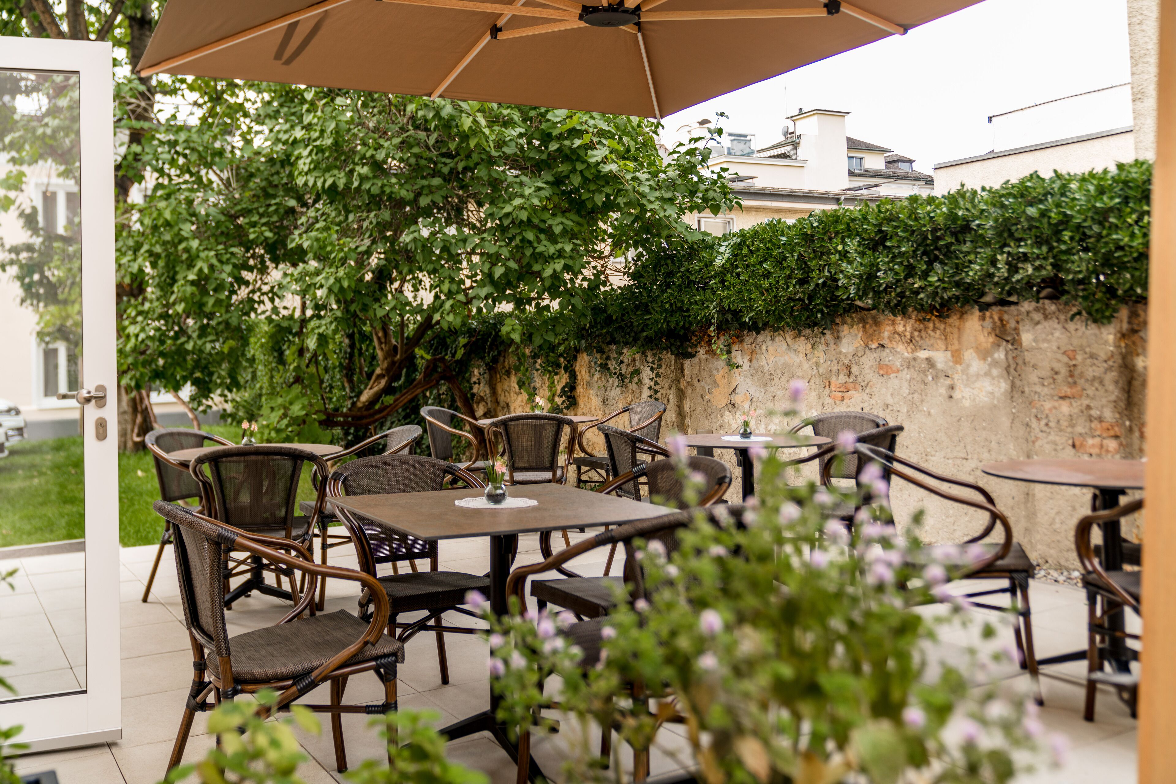 An outdoor patio with wicker furniture and a large umbrella, surrounded by greenery and a stone wall.