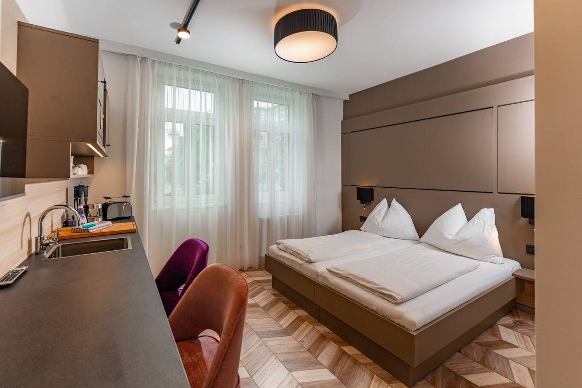 A modern hotel room with a bed, a desk with a chair, a sink, and a window with curtains.