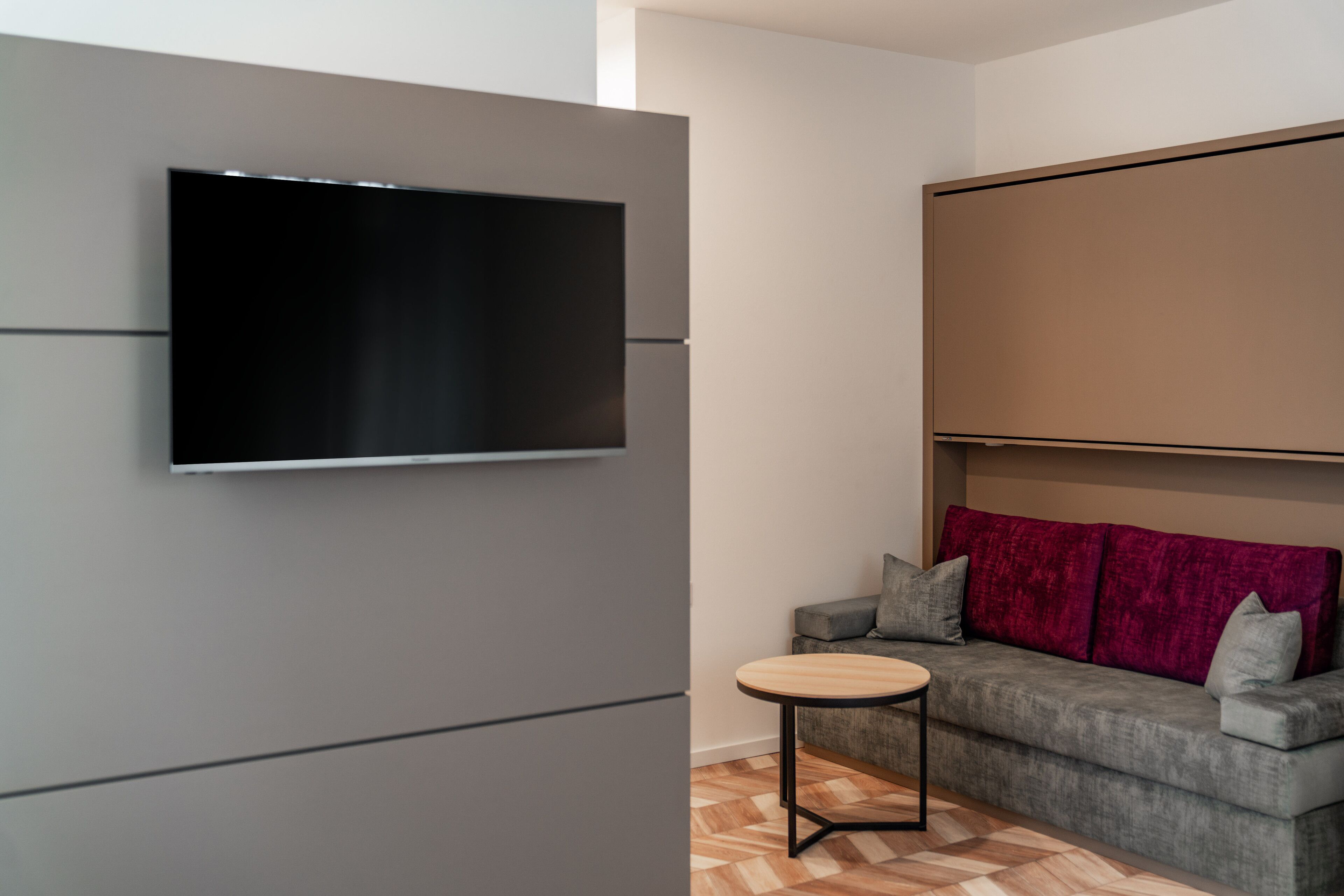 Uma sala de estar moderna com uma televisão de tela plana fixada em uma parede cinza, um sofá cinza com almofadas vermelhas e uma pequena mesa redonda.