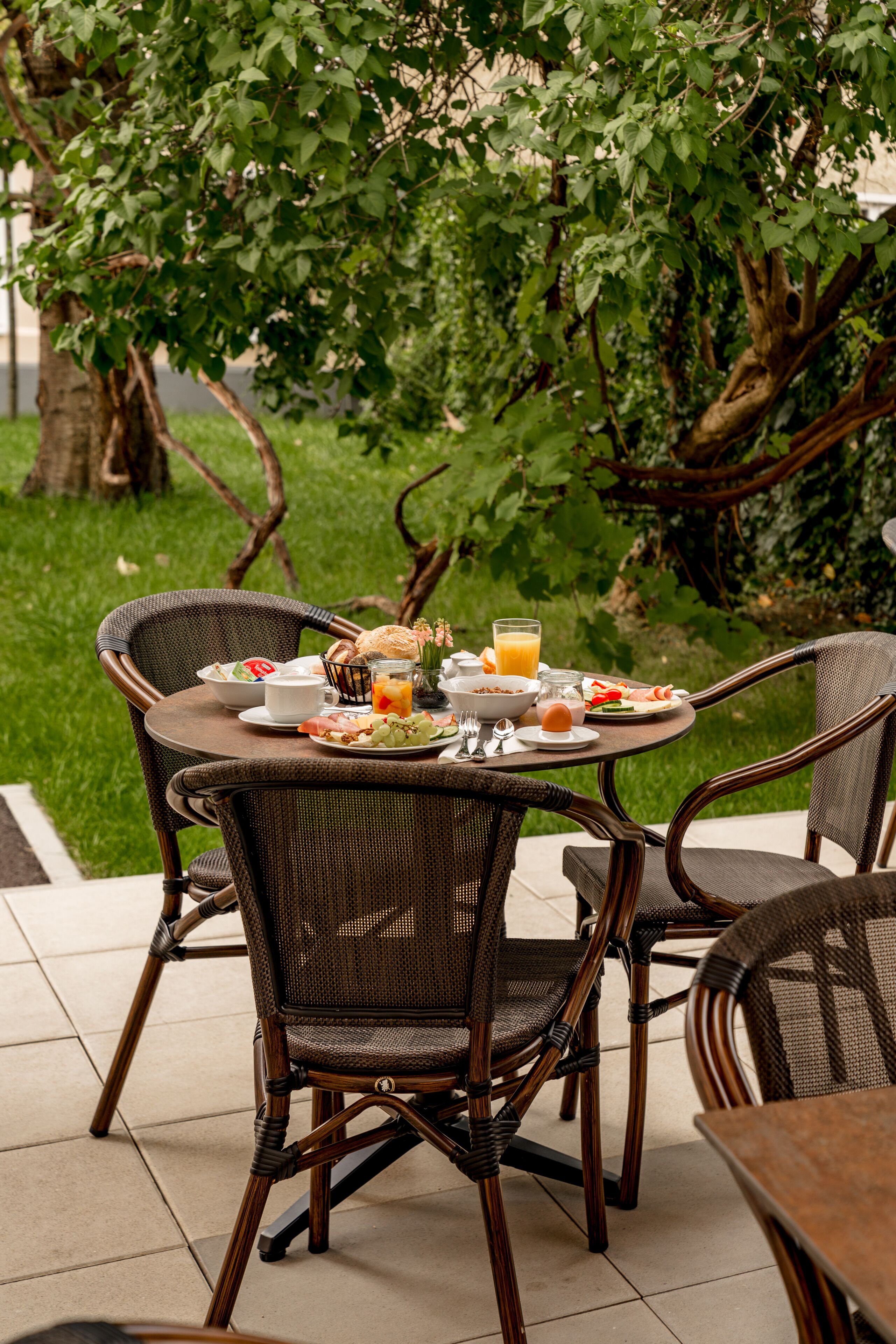 Ein runder Tisch mit Korbstühlen, Frühstücksleckereien und einem Glas Orangensaft auf einer Terrasse, im Hintergrund ein Baum und Rasen.