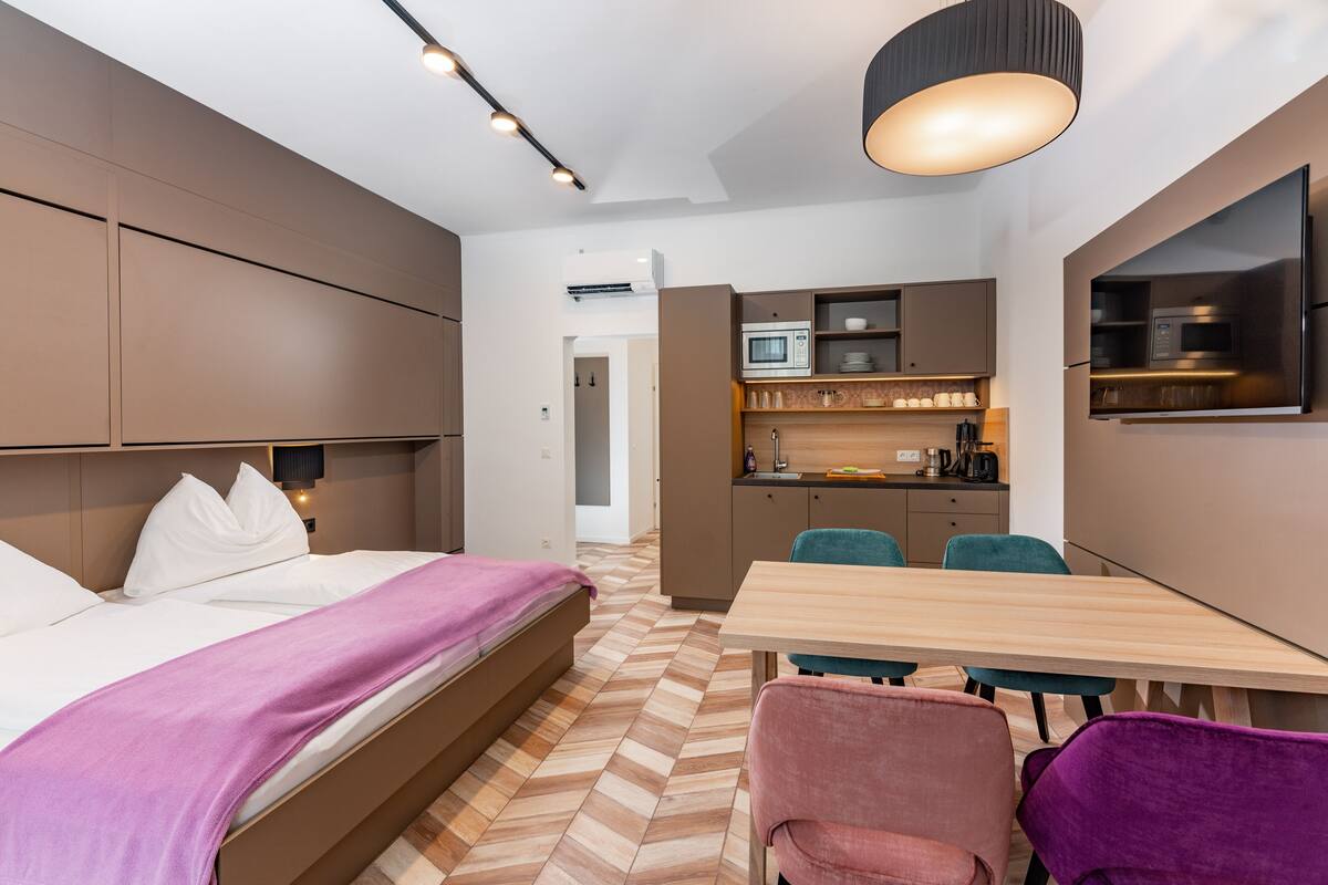 A modern hotel room with a bed, a dining table, and a kitchenette.
