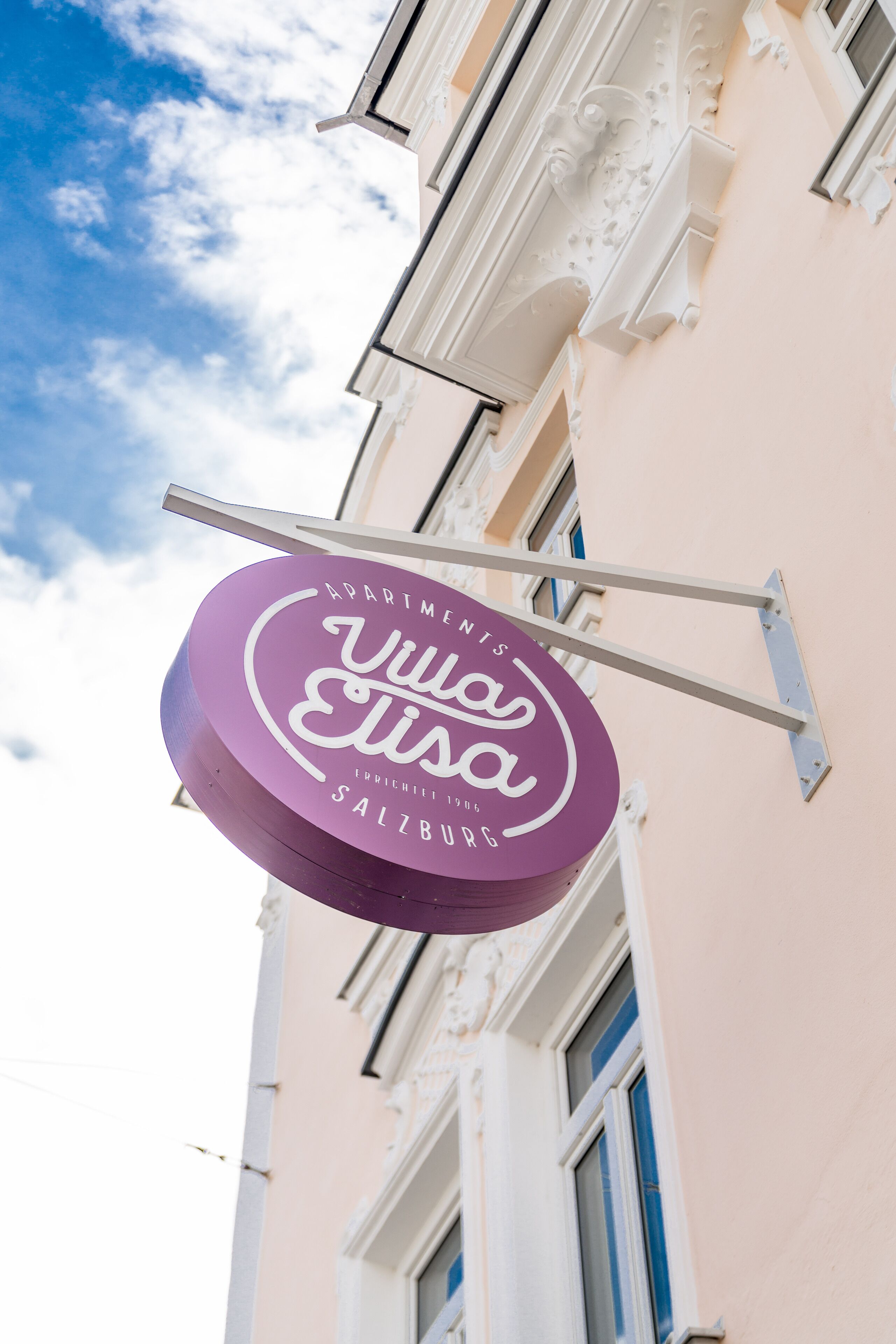 A sign for "Villa Elisa Apartments" with a clear view of the building's facade and a partly cloudy sky.