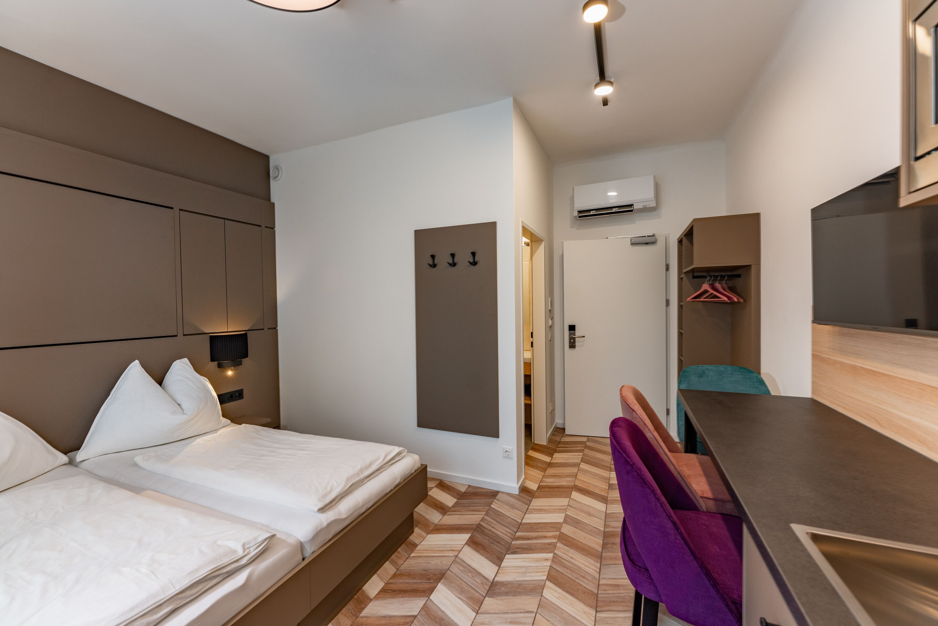 A hotel room with a bed, a desk, a chair, and an air conditioning unit.