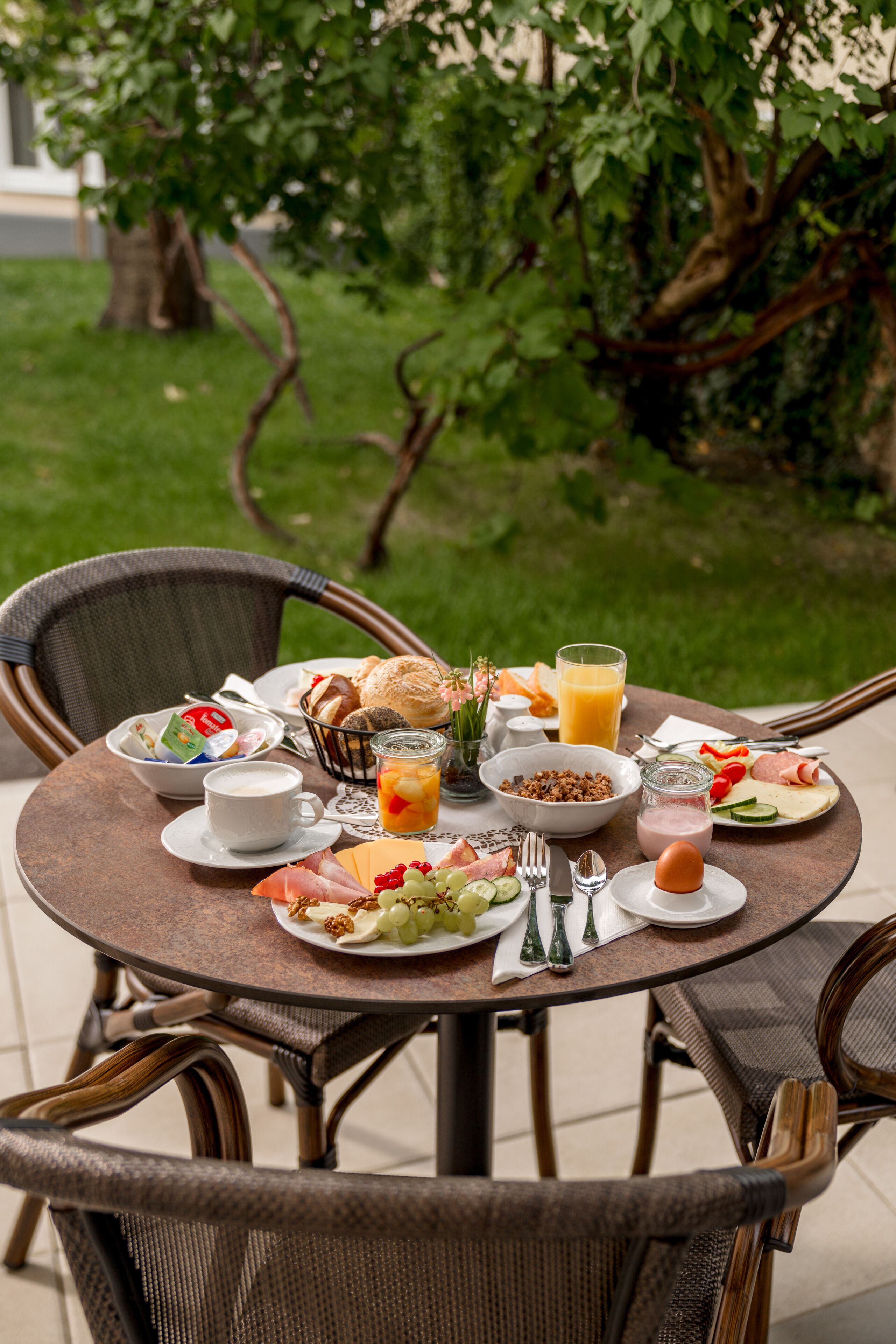 Una mesa redonda con alimentos para el desayuno, como huevos, pan y jugo, ubicada al aire libre con sillas y un árbol de fondo.