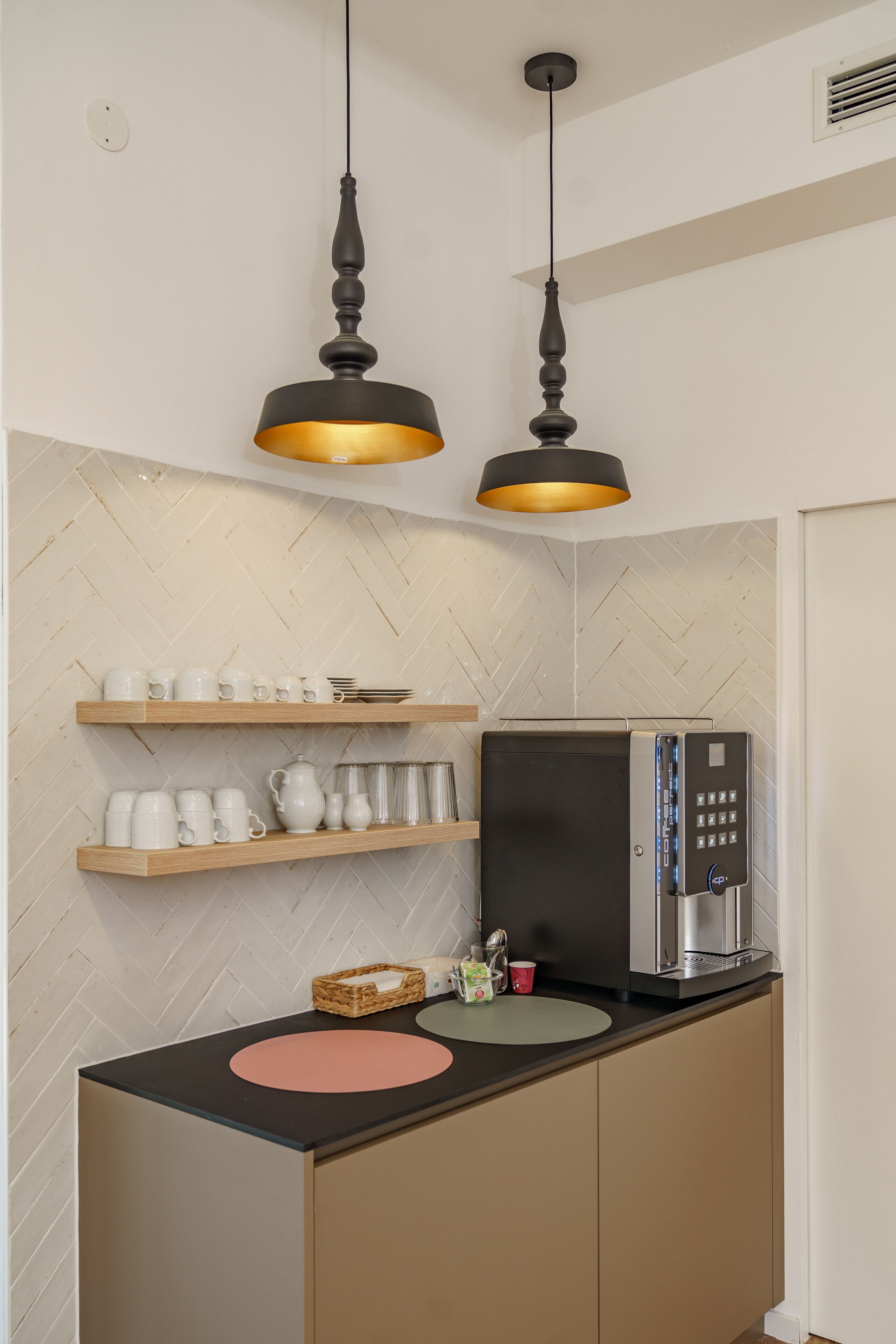 Una kitchenette con dos lámparas colgantes, estantes de madera con utensilios de cocina, un microondas y un refrigerador pequeño.