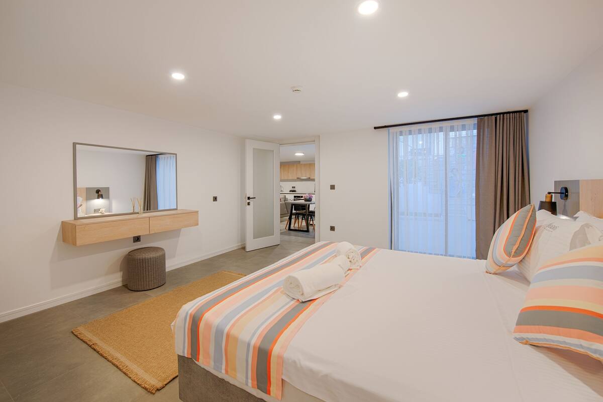 Una habitación de hotel moderna con una cama grande, una mesita de noche, un espejo y vistas a la cocina y al comedor por una puerta abierta.