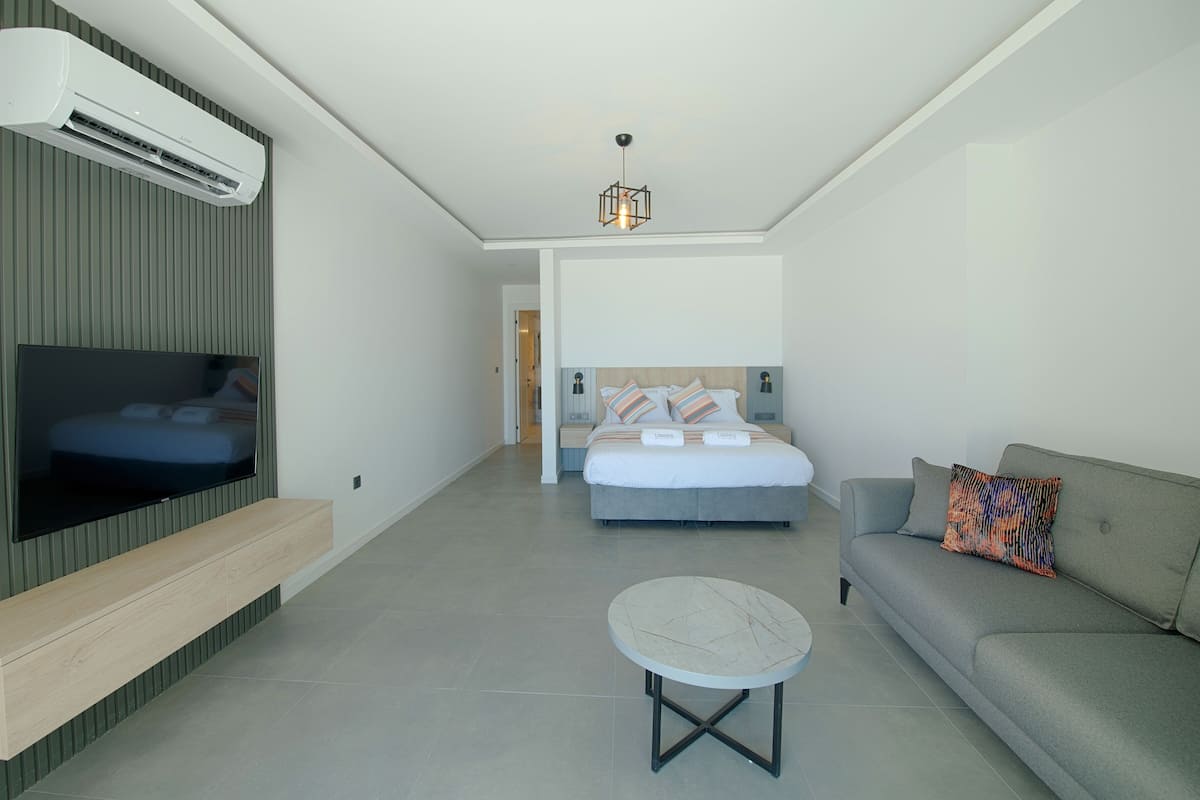 Una habitación de hotel moderna con un televisor de pantalla plana montado en la pared, una cama con cabecera de madera, un sofá gris, una mesita redonda pequeña y una lámpara colgante.