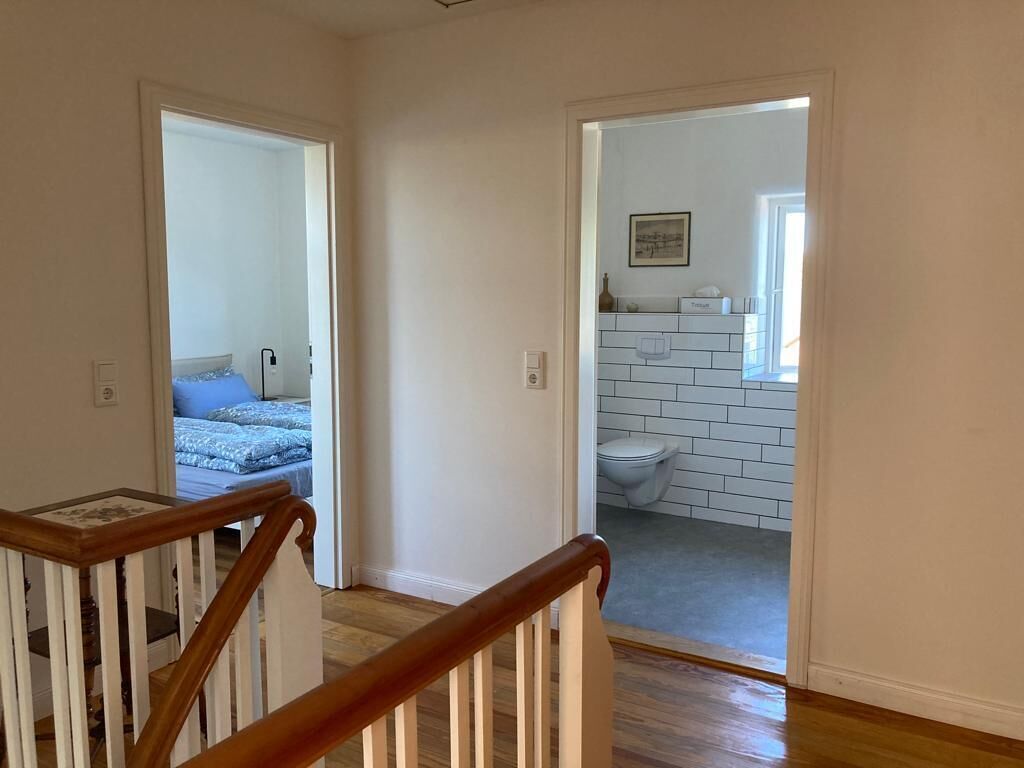Un couloir menant à une chambre et à une salle de bain, le tout carrelé de blanc, avec un tableau encadré accroché au mur.