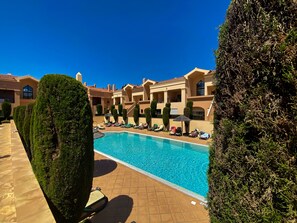 Ein Poolbereich mit Liegestühlen und einem klaren, blauen Pool, umgeben von ordentlich geschnittenen Hecken und einem Gebäude mit einem Ziegeldach.