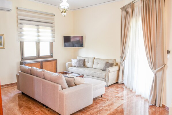 Un soggiorno con un divano beige, un divano più piccolo, una televisione e una finestra con persiane.
