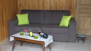 Un salón con un sofá gris, dos almohadas verdes, una mesa de centro de madera con un mantel blanco y una mesita auxiliar con libros.